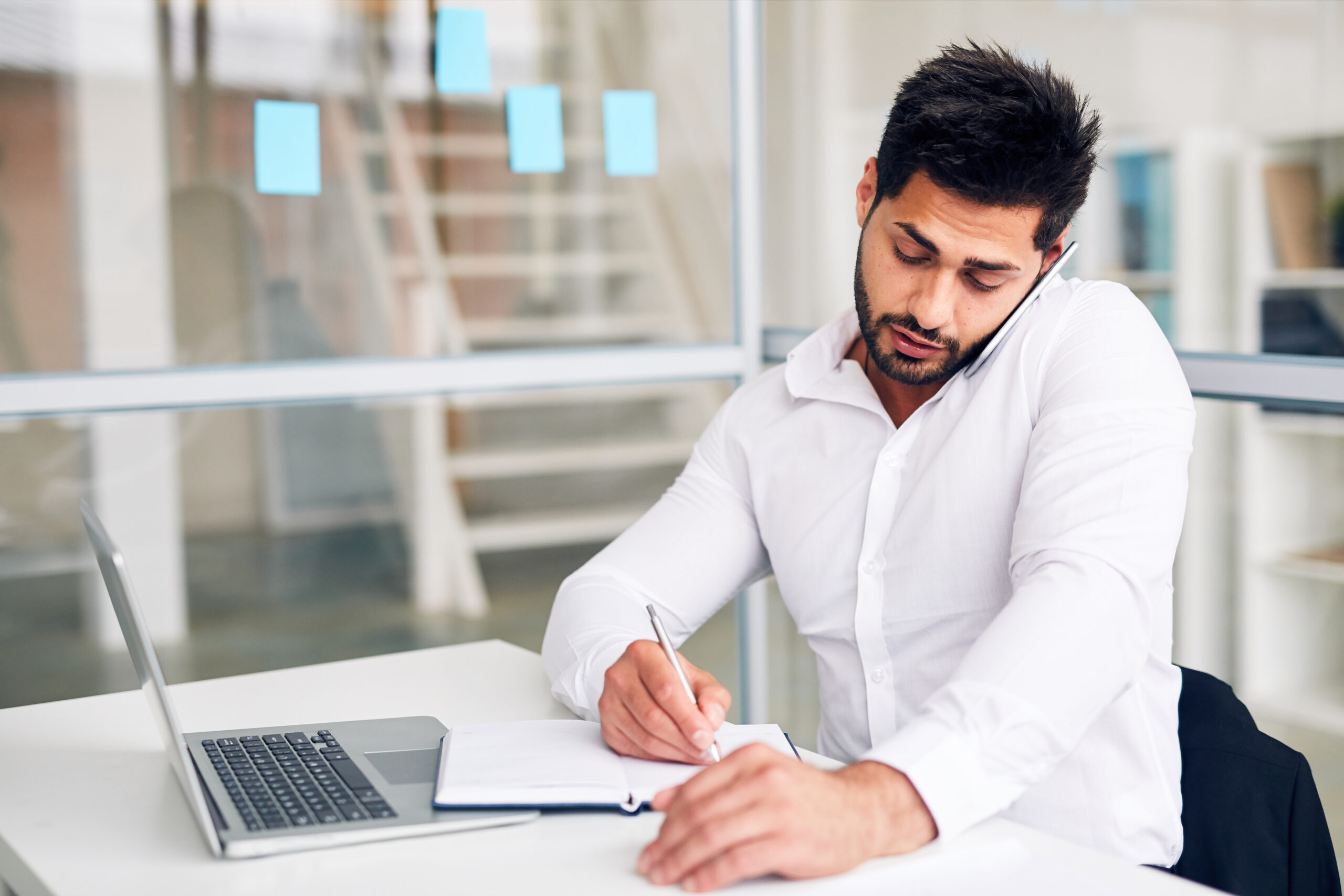 Multi-tasking office worker making notes in notebook while consulting someone by smartphone